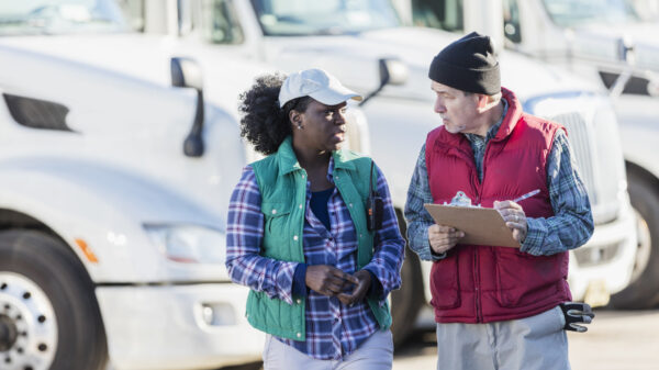 Two multi-ethnic truck drivers, a senior Hispanic man in his 60s and a mid-adult African-American woman in her 30s, standing in front of a fleet of semi-trucks or tractor-trailers. The man is holding a clipboard and they are conversing, looking at each other face to face.