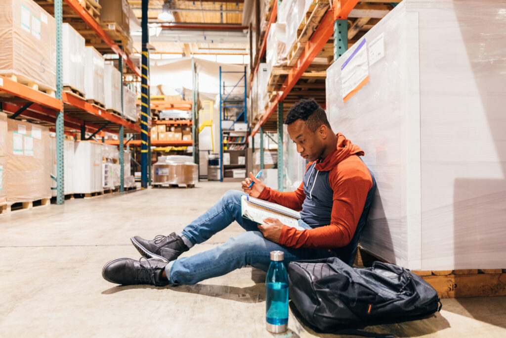 A male printing press and warehouse worker finds some spare time to study while on break and further his education as a university and college student. He is studying from a textbook, writing notes.