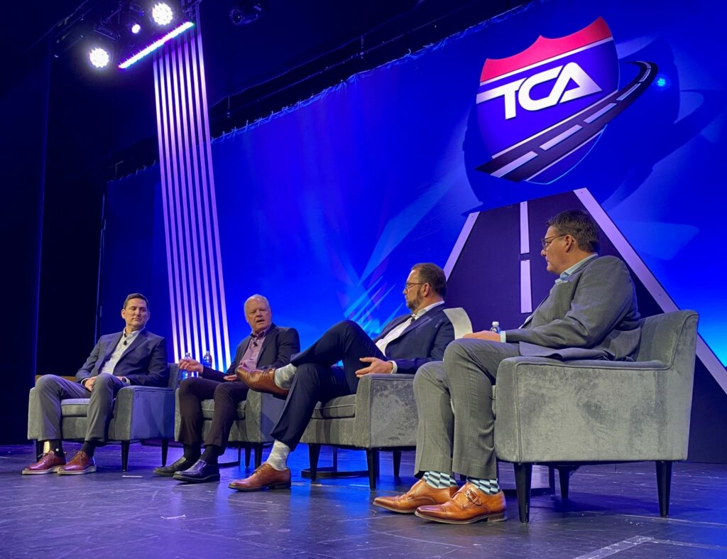 Men seated on a stage during a TCA discussion.