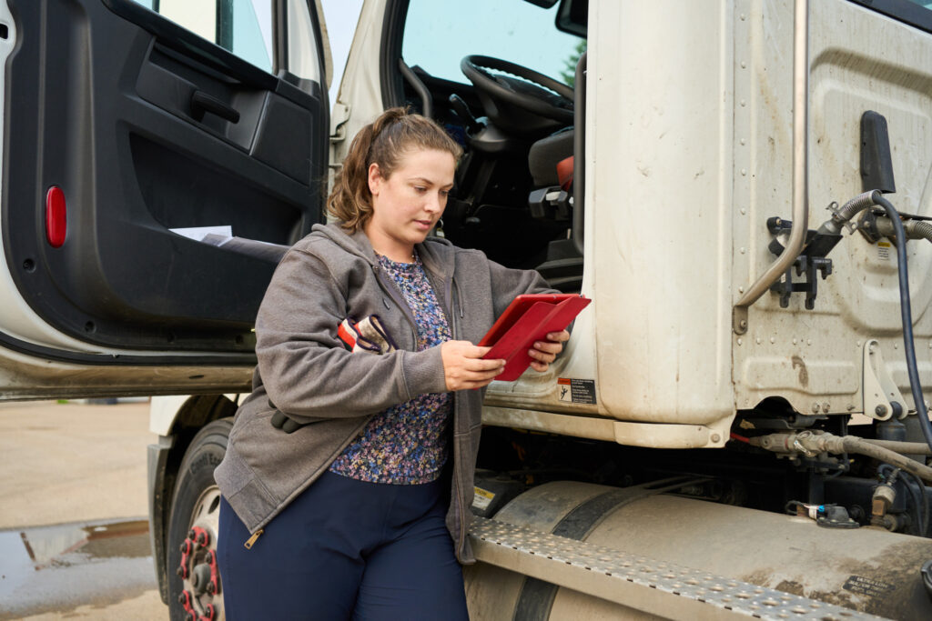 Young female truck driver leaning on her truck cabin at highway stop and looking at routes on a digital tablet